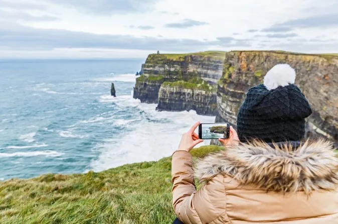 Une visiteuse en bonnet à pompon et manteau à col de fourrure qui photographie les falaises avec un téléphone