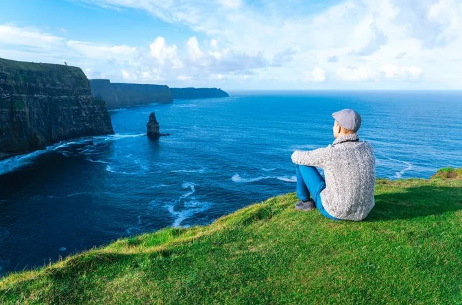 A man in a cap sitting on the grass looking out over the cliffs and a sea stack